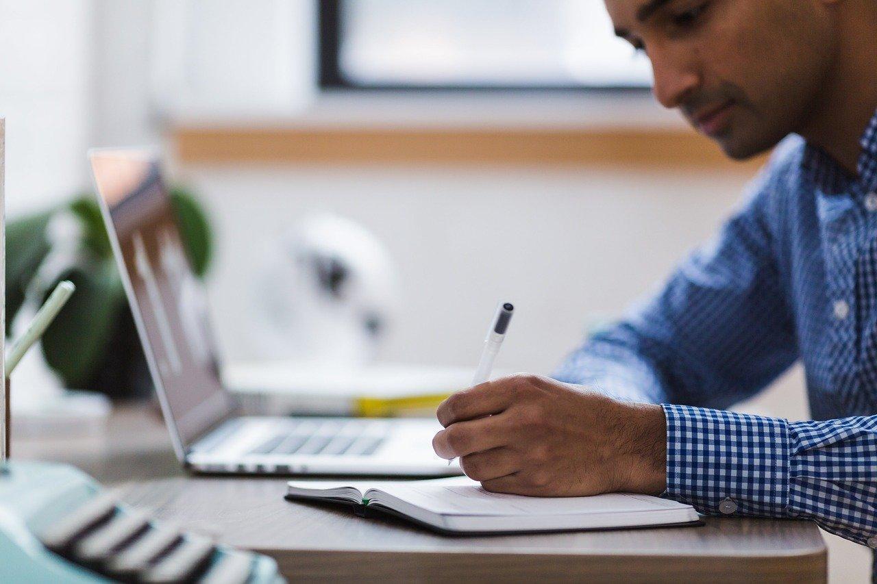 Man writes in a notebook next to a laptop